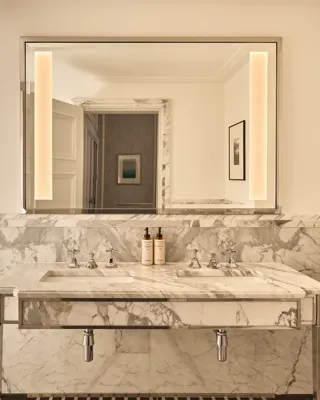 Marble double vanity with twin sinks, illuminated mirror and stone-clad walls, viewed through an open doorway into the bathroom.
