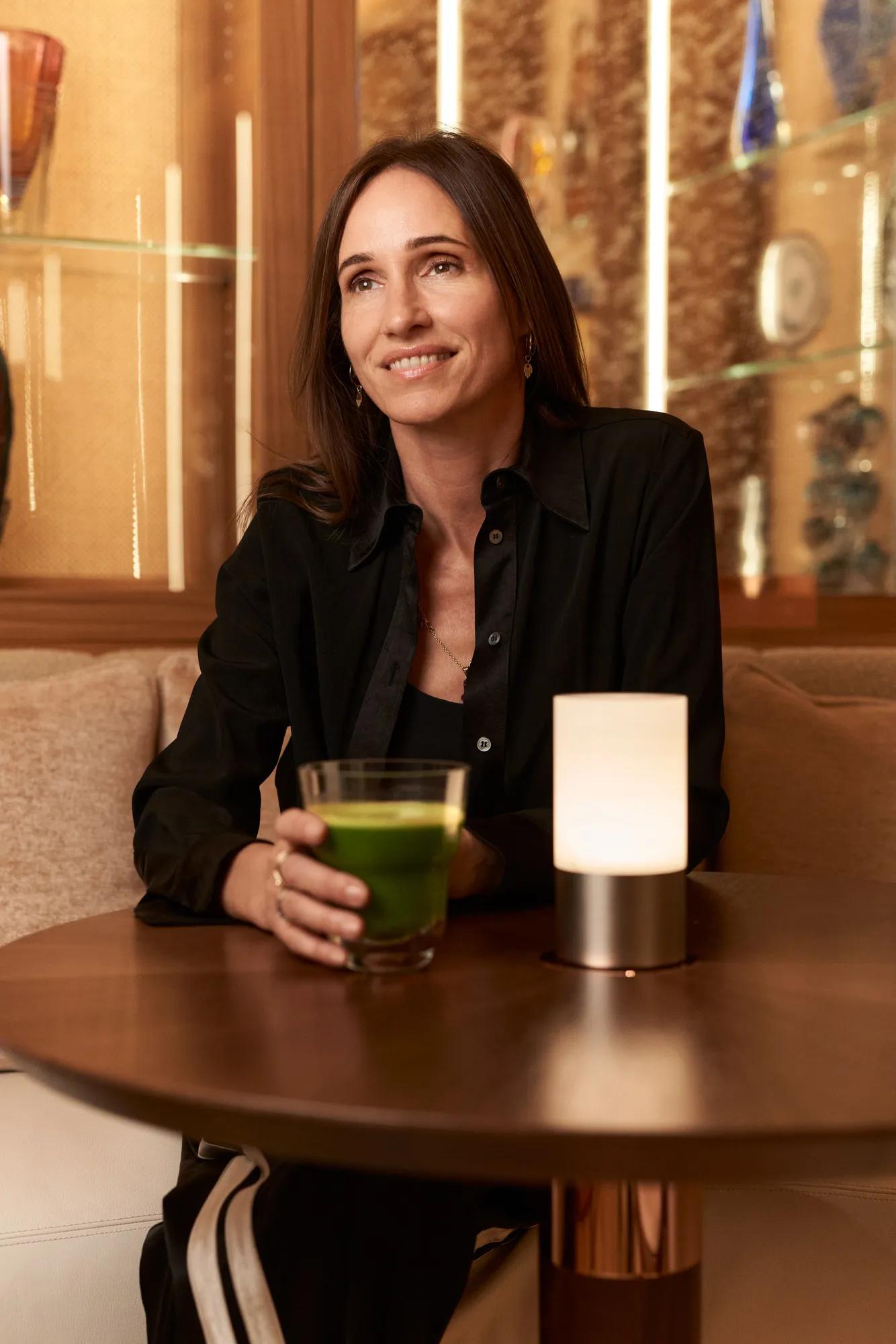 Woman seated at café table holding green juice, smiling, with small lamp and glass display shelves behind.