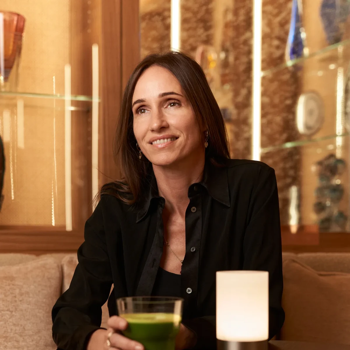Woman seated at café table holding green juice, smiling, with small lamp and glass display shelves behind.