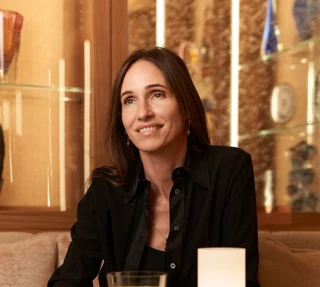 Woman seated at café table holding green juice, smiling, with small lamp and glass display shelves behind.