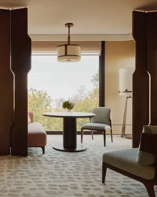 Hotel suite with bedroom and seating area, featuring wood panelling, round table, armchairs and large window with tree views.
