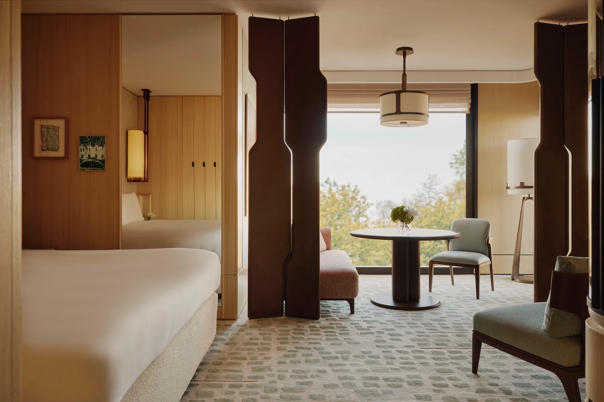 Hotel suite with bedroom and seating area, featuring wood panelling, round table, armchairs and large window with tree views.