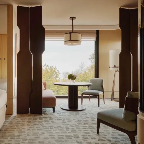 Hotel suite with bedroom and seating area, featuring wood panelling, round table, armchairs and large window with tree views.