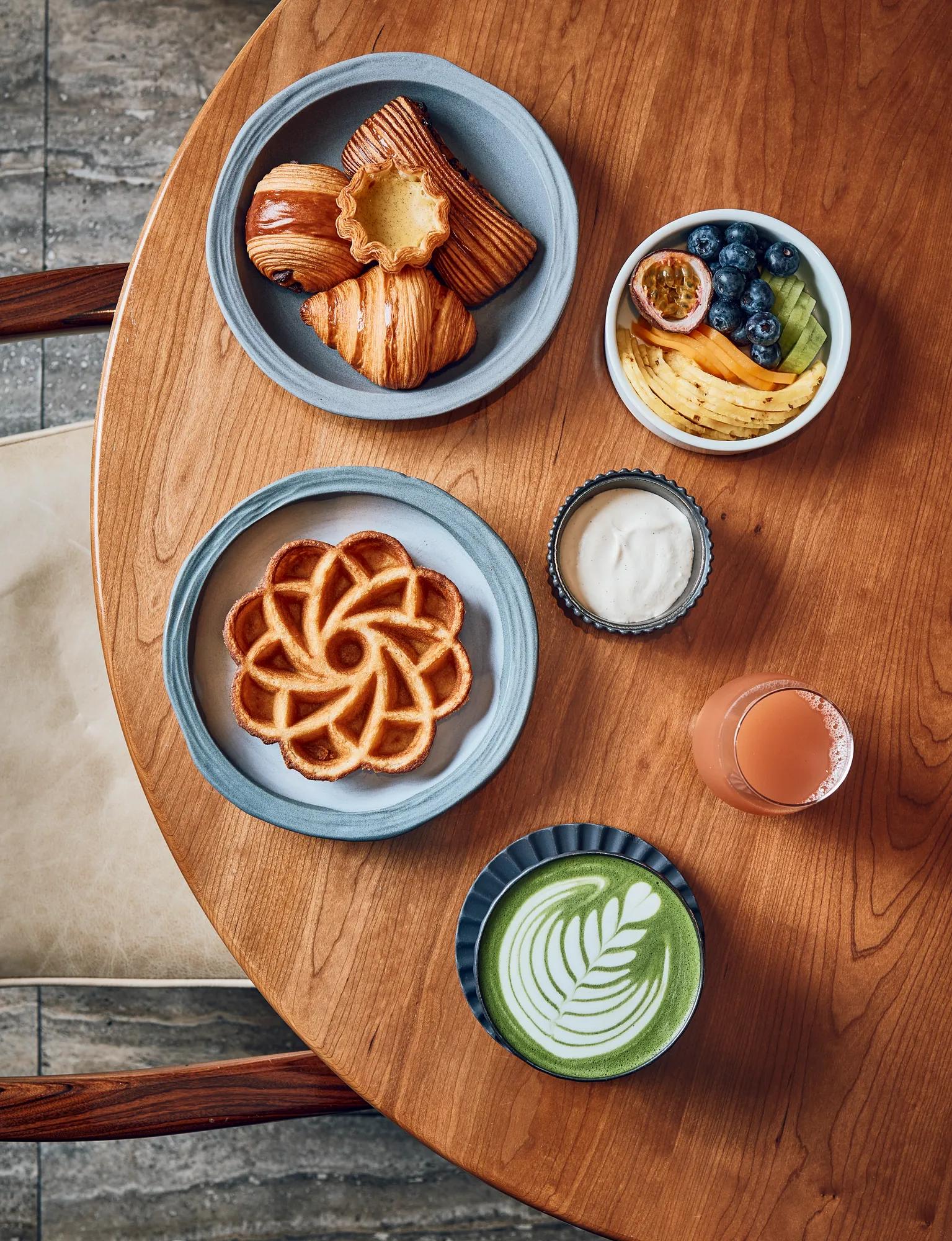 Breakfast spread with pastries, waffle, fruit bowl, matcha latte, juice, and yogurt on wooden table