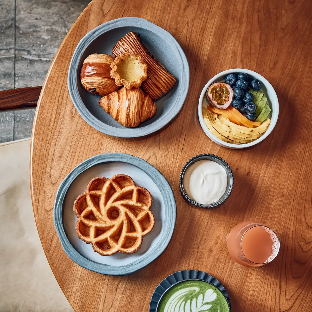 Breakfast spread with pastries, waffle, fruit bowl, matcha latte, juice, and yogurt on wooden table