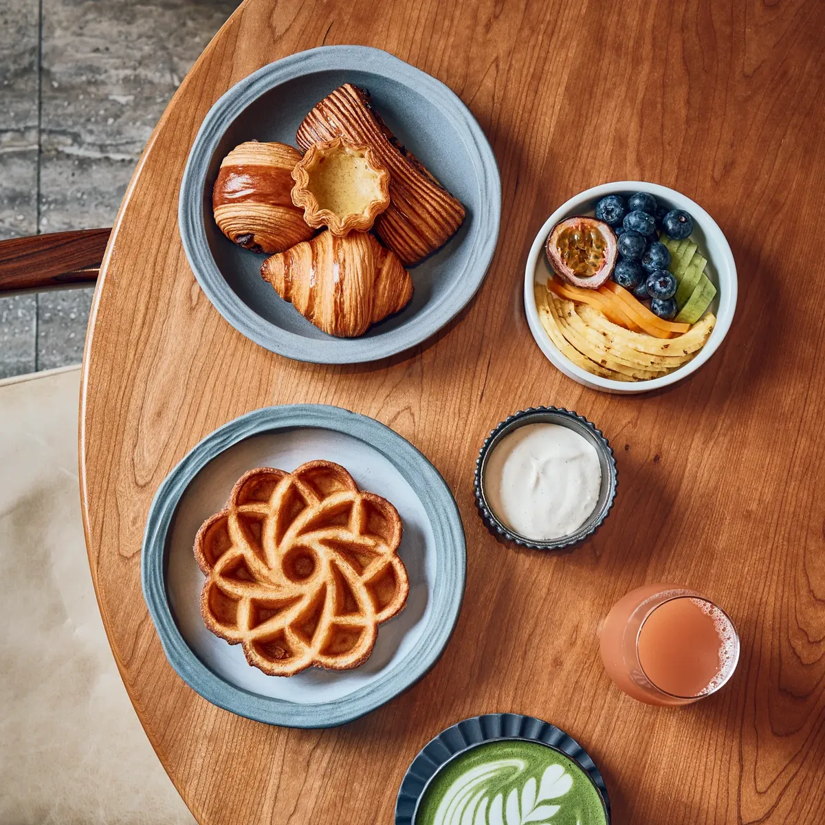 Breakfast spread with pastries, waffle, fruit bowl, matcha latte, juice, and yogurt on wooden table