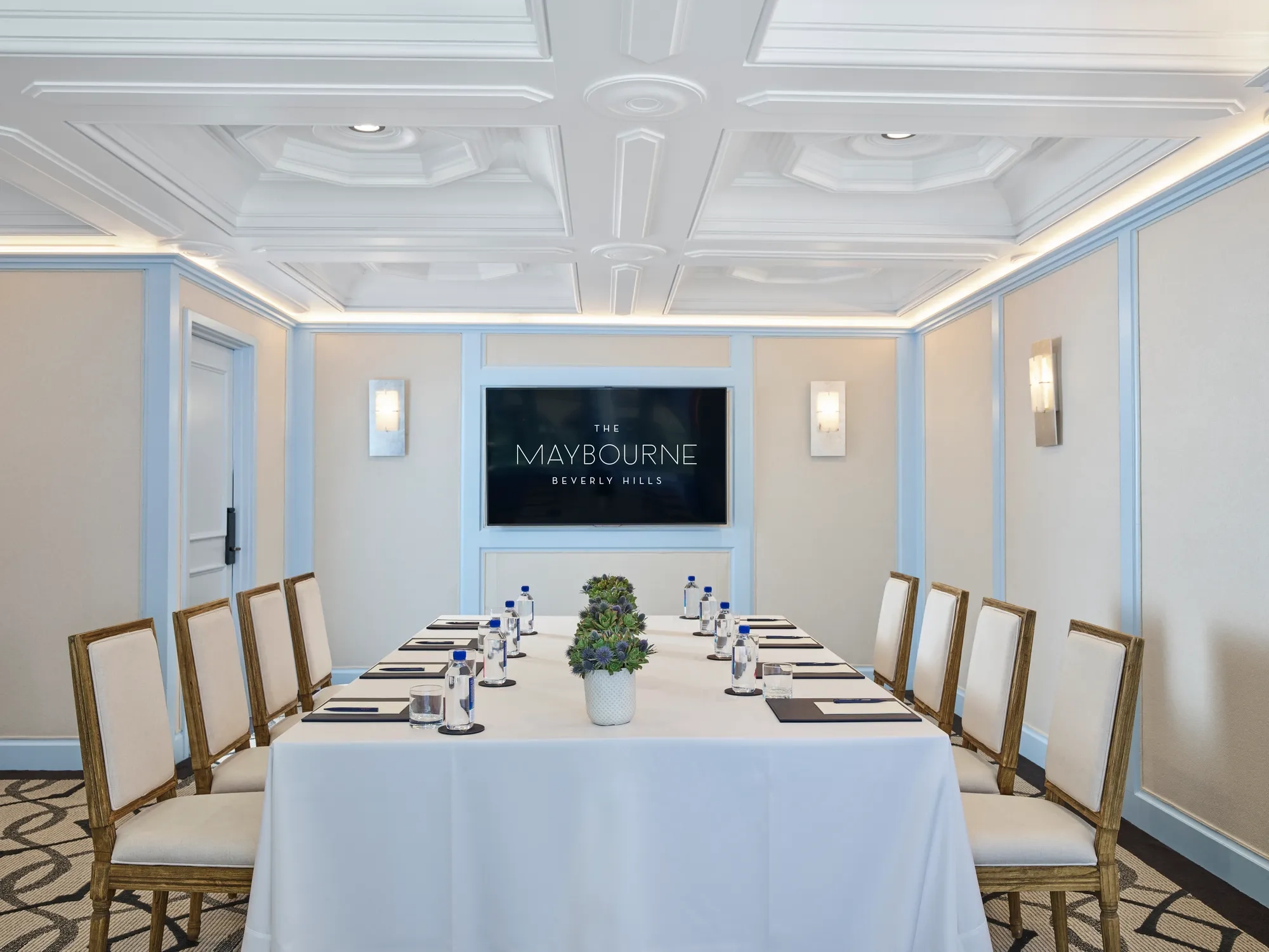Wilshire Room boardroom setup with U-shaped table, neutral chairs, water and notepads, and a wall display screen