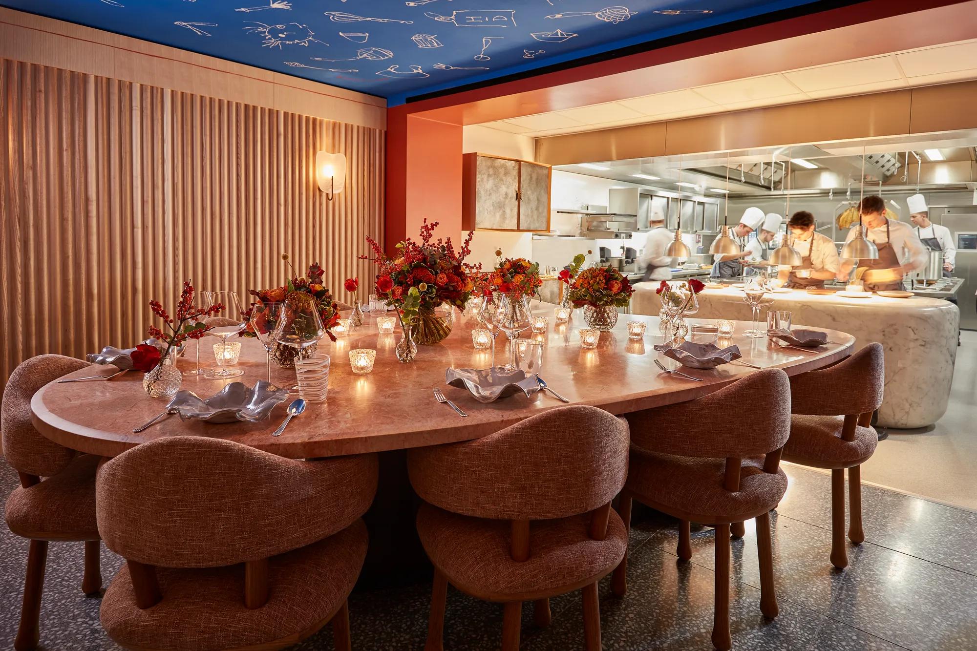 Large round dining table set with flowers and candles, facing an open kitchen where chefs are cooking