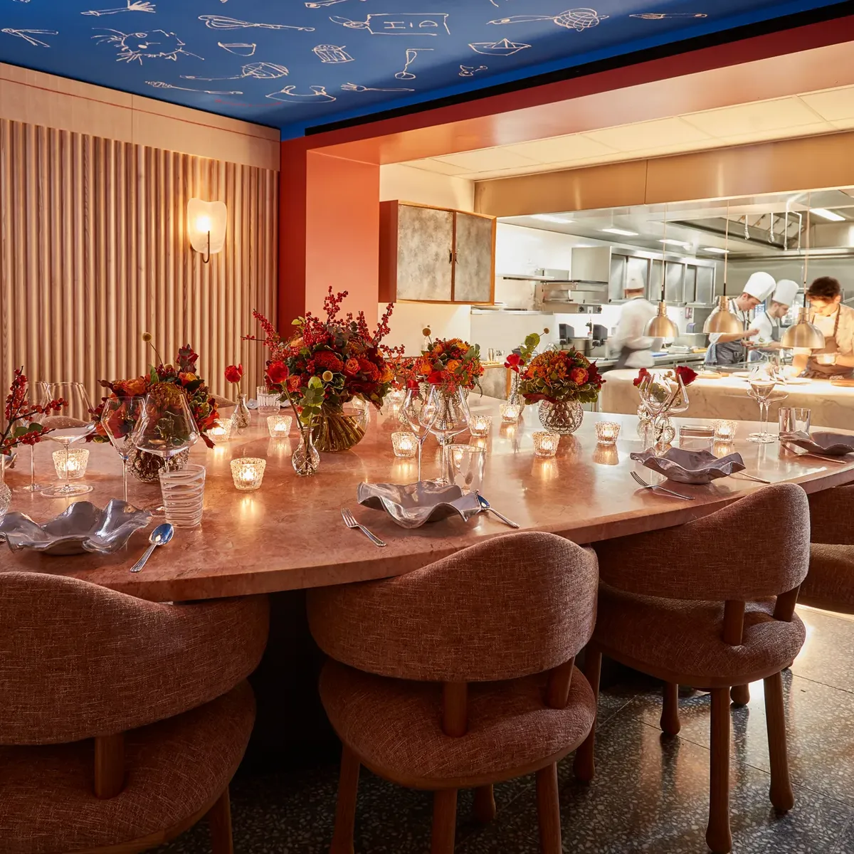 Large round dining table set with flowers and candles, facing an open kitchen where chefs are cooking
