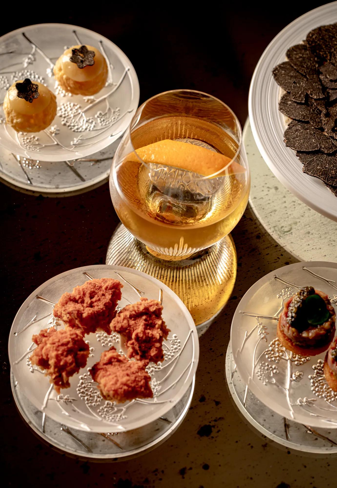 Glass of amber-coloured drink surrounded by elegant canapé plates and shaved truffles.