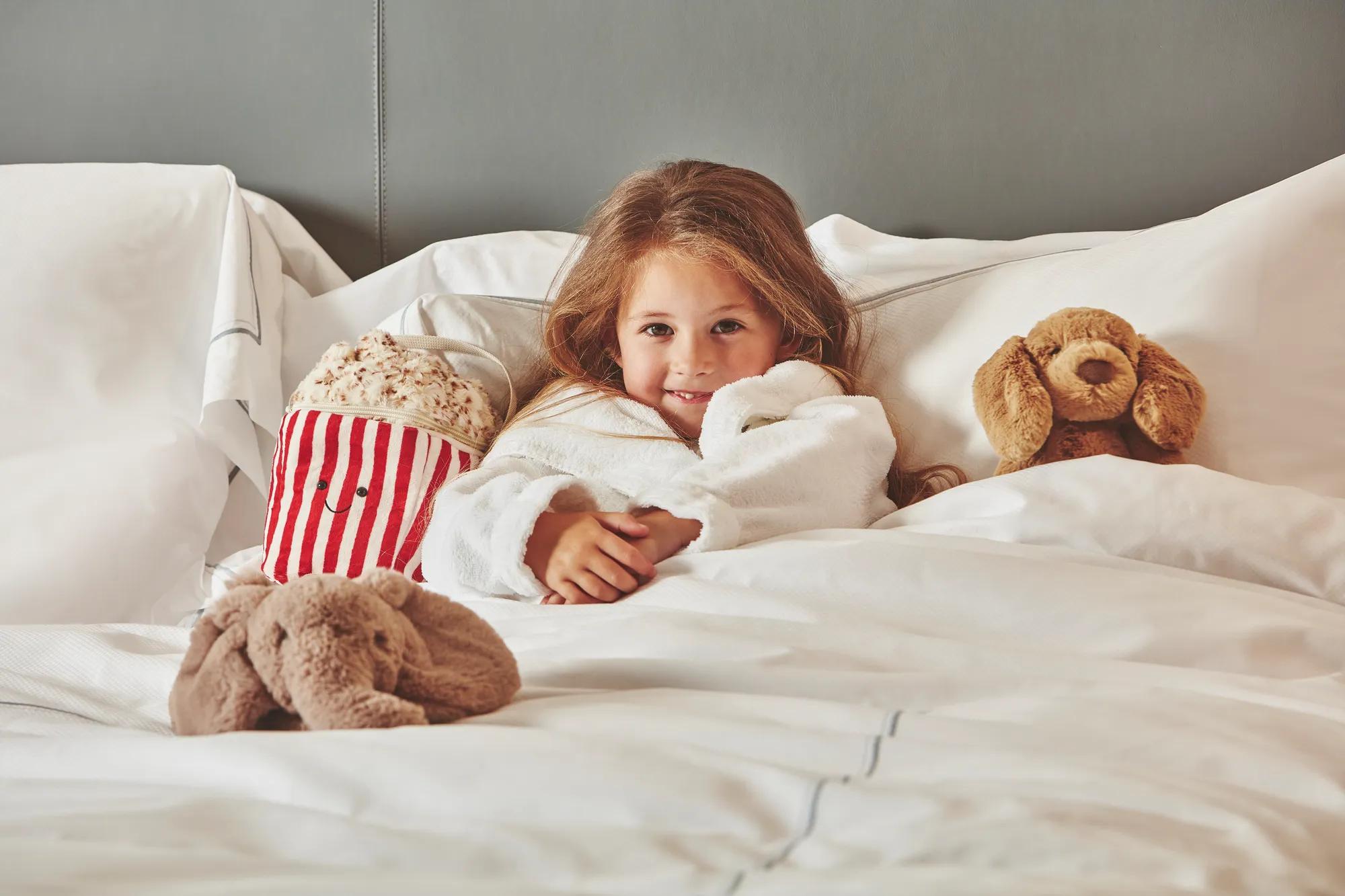 Child relaxing in a hotel bed with plush toys and a striped popcorn tub, wrapped in a white robe.