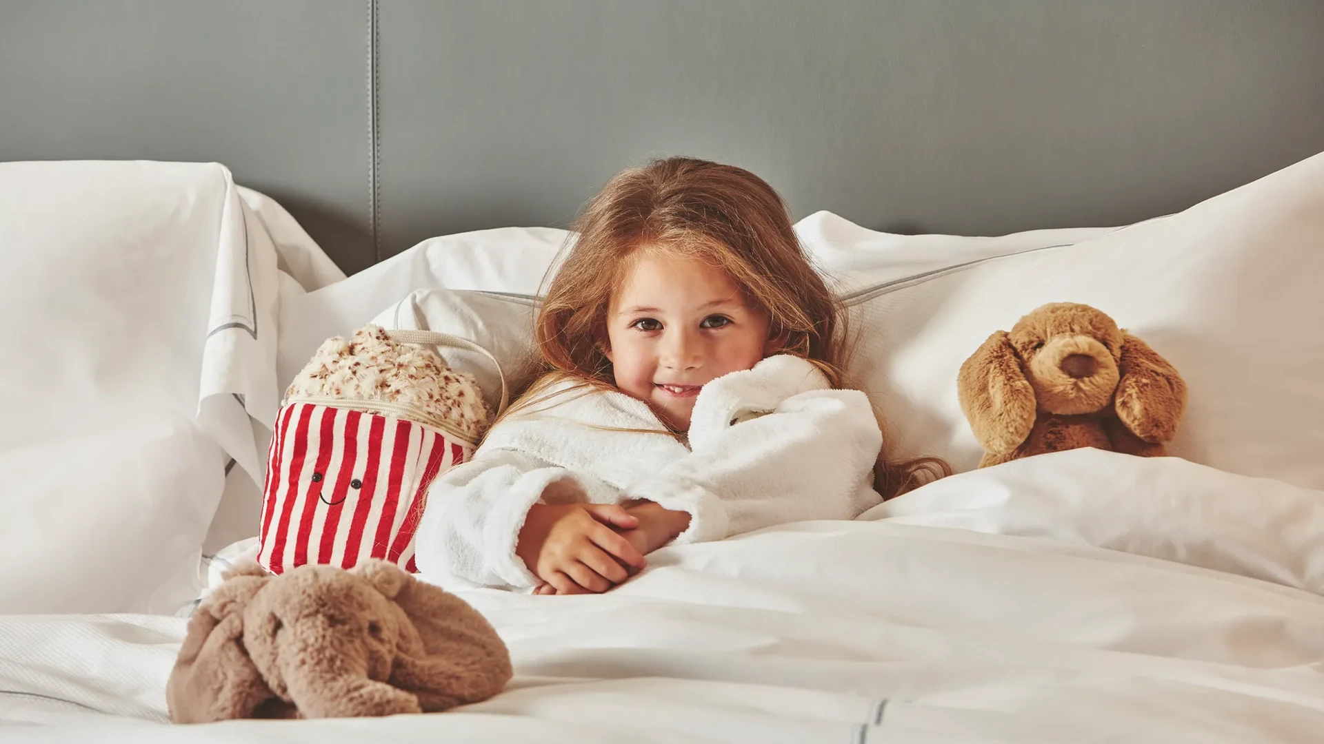 Child relaxing in a hotel bed with plush toys and a striped popcorn tub, wrapped in a white robe.