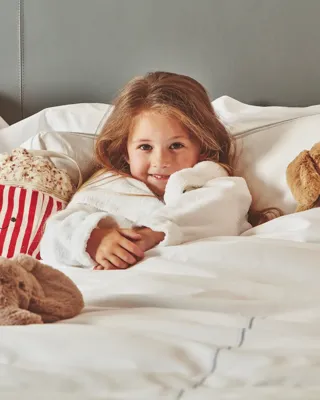 Child relaxing in a hotel bed with plush toys and a striped popcorn tub, wrapped in a white robe.