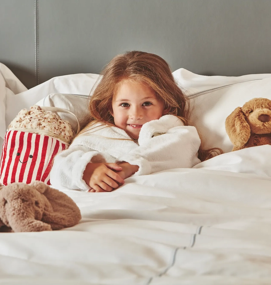 Child relaxing in a hotel bed with plush toys and a striped popcorn tub, wrapped in a white robe.