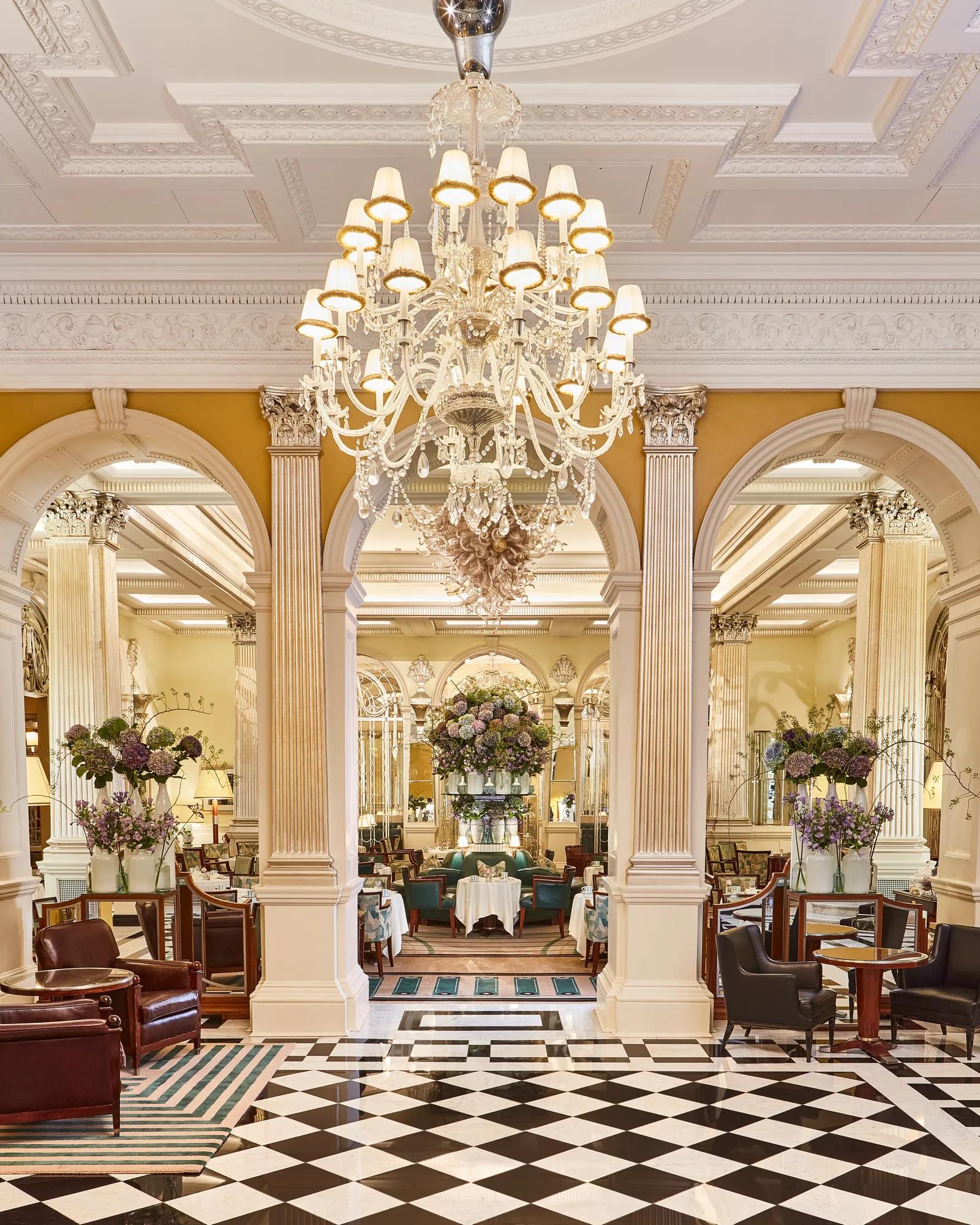 An elegant dining room with grand chandeliers, marble columns, and checkerboard flooring, featuring tables set with white linens and floral arrangements of purple and green blooms beneath ornate high ceilings.