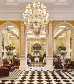 An elegant dining room with grand chandeliers, marble columns, and checkerboard flooring, featuring tables set with white linens and floral arrangements of purple and green blooms beneath ornate high ceilings.