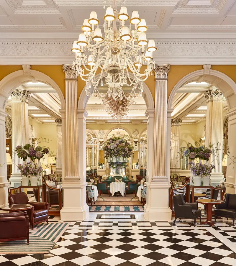 An elegant dining room with grand chandeliers, marble columns, and checkerboard flooring, featuring tables set with white linens and floral arrangements of purple and green blooms beneath ornate high ceilings.
