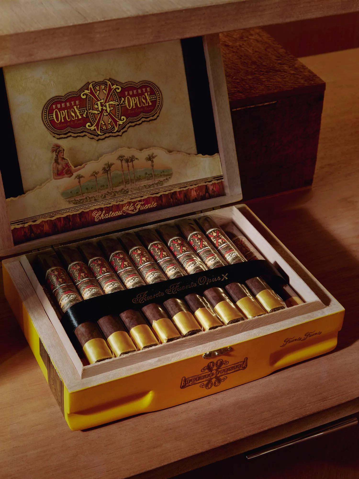 Open yellow Fuente Fuente OpusX cigar box on a wood shelf, rows of banded cigars inside, decorative lid art above.