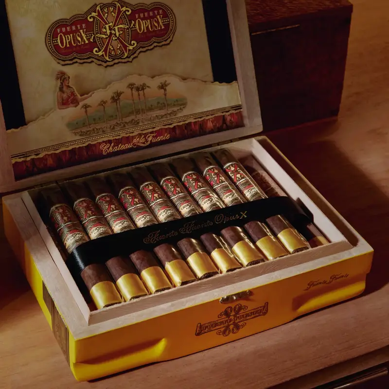 Open yellow Fuente Fuente OpusX cigar box on a wood shelf, rows of banded cigars inside, decorative lid art above.