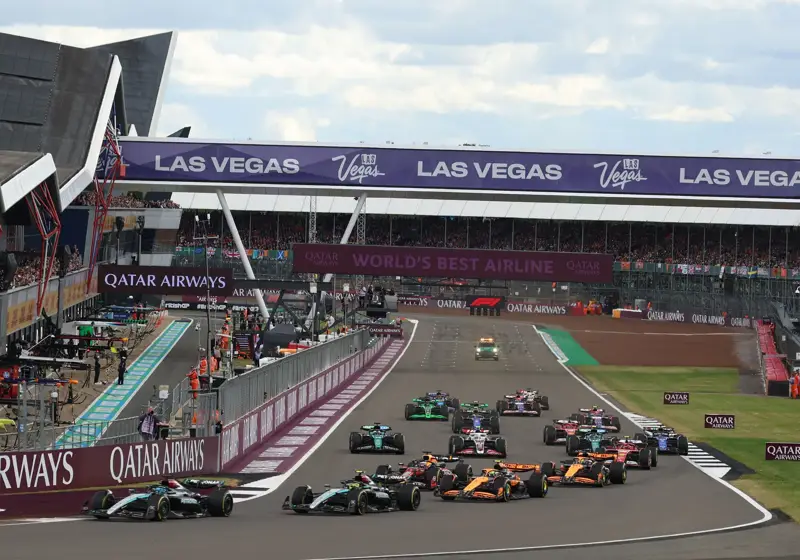 Formula 1 cars race tightly into corner beneath Las Vegas banner, crowded grandstands and pit lane visible.