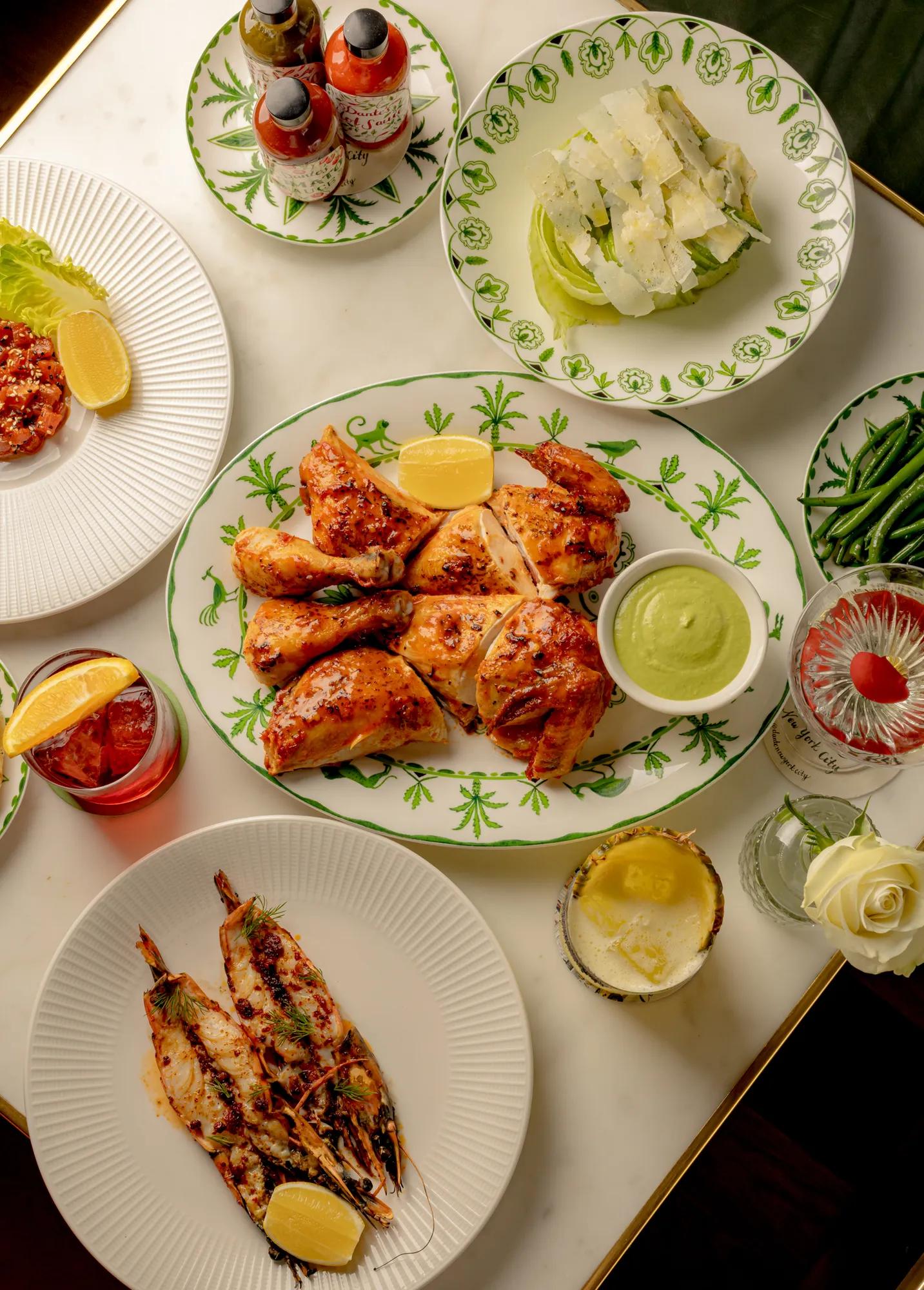Roast chicken platter with green dipping sauce, grilled prawns, Caesar salad, cocktails, and side dishes arranged on a marble dining table.