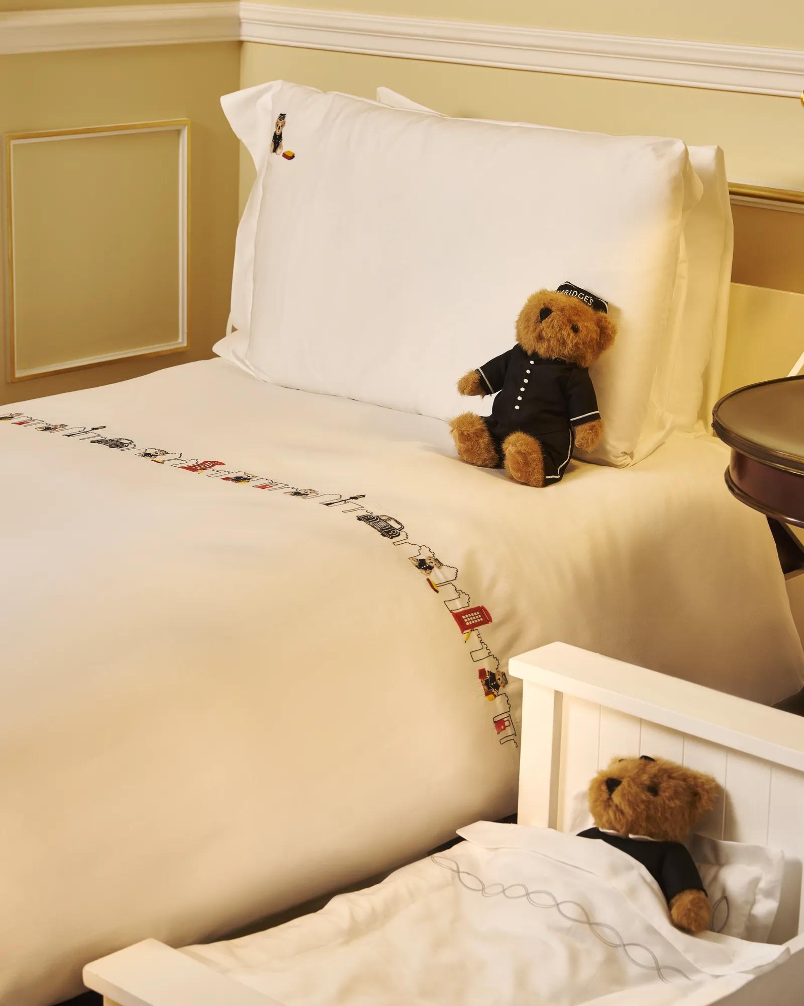 Bed with white linens, embroidered trim, and teddy bear in uniform on pillow; second teddy bear rests in small child’s cot beside bed.