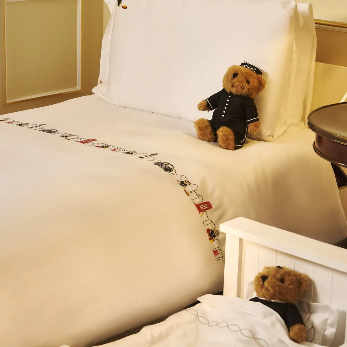 Bed with white linens, embroidered trim, and teddy bear in uniform on pillow; second teddy bear rests in small child’s cot beside bed.