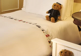 Bed with white linens, embroidered trim, and teddy bear in uniform on pillow; second teddy bear rests in small child’s cot beside bed.