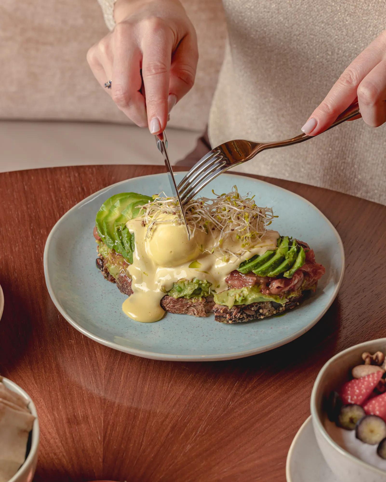 Hands cutting eggs Benedict with avocado on toast, topped with hollandaise sauce, served on a ceramic plate.