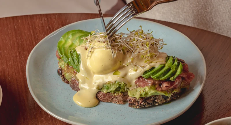 Hands cutting eggs Benedict with avocado on toast, topped with hollandaise sauce, served on a ceramic plate.