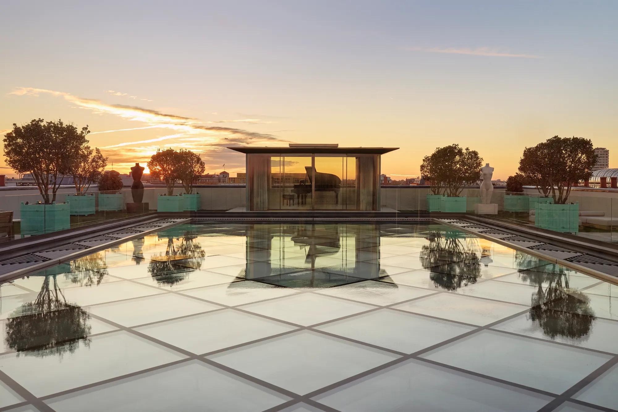 Glass-roof terrace at Claridge’s with a pavilion housing a grand piano, surrounded by trees and sculptures at sunset