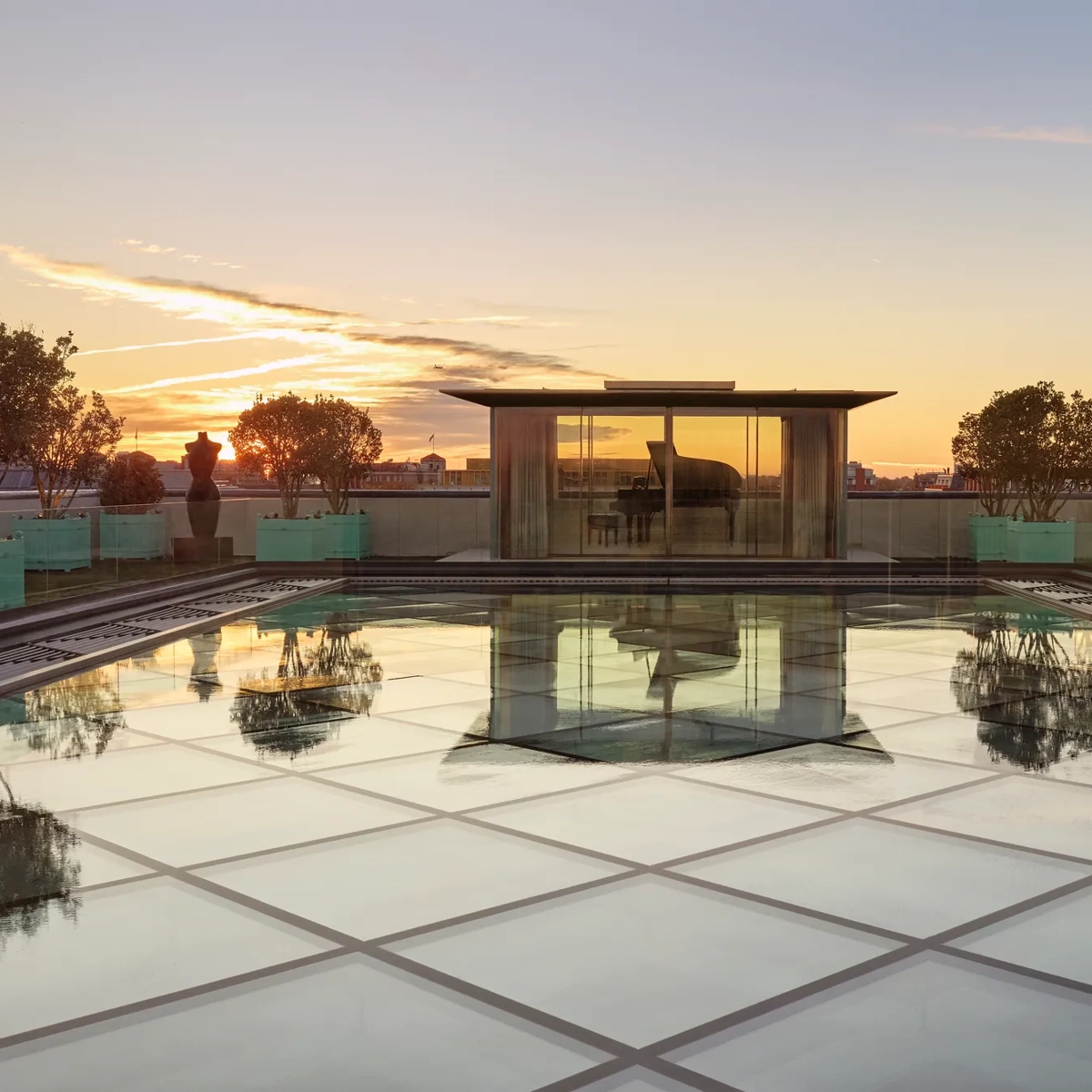 Glass-roof terrace at Claridge’s with a pavilion housing a grand piano, surrounded by trees and sculptures at sunset.