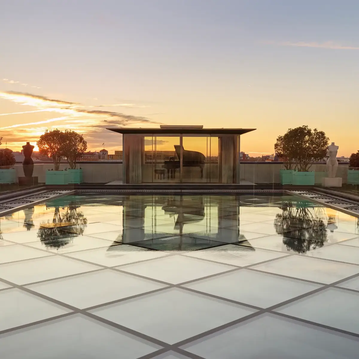 Glass-roof terrace at Claridge’s with a pavilion housing a grand piano, surrounded by trees and sculptures at sunset.