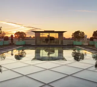 Glass-roof terrace at Claridge’s with a pavilion housing a grand piano, surrounded by trees and sculptures at sunset.