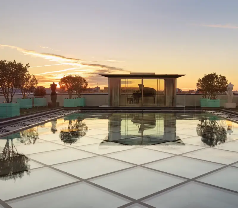 Glass-roof terrace at Claridge’s with a pavilion housing a grand piano, surrounded by trees and sculptures at sunset.