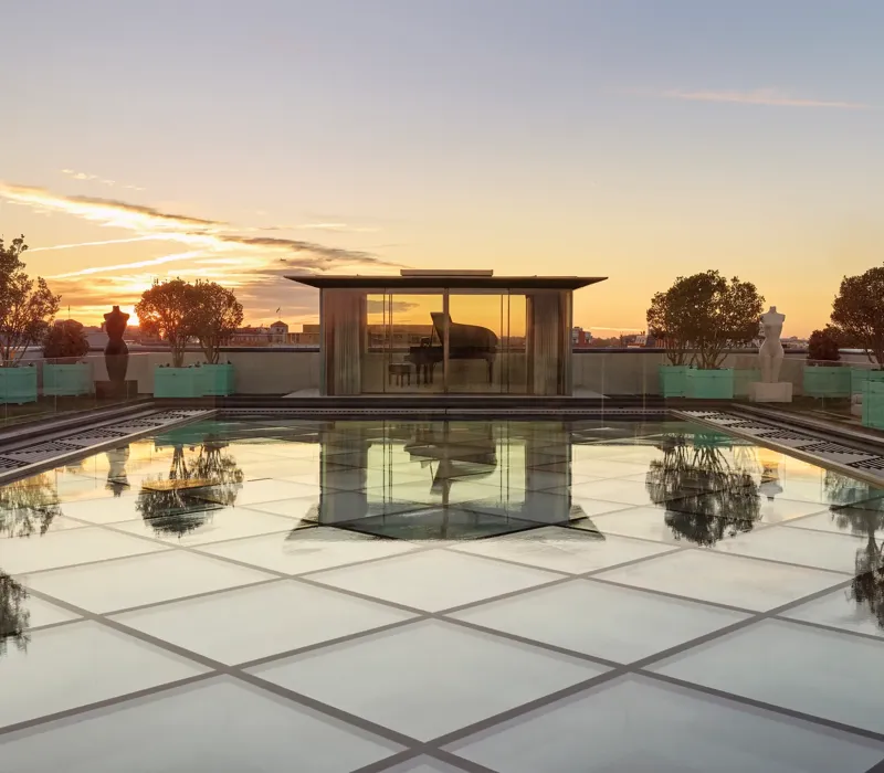 Glass-roof terrace at Claridge’s with a pavilion housing a grand piano, surrounded by trees and sculptures at sunset.