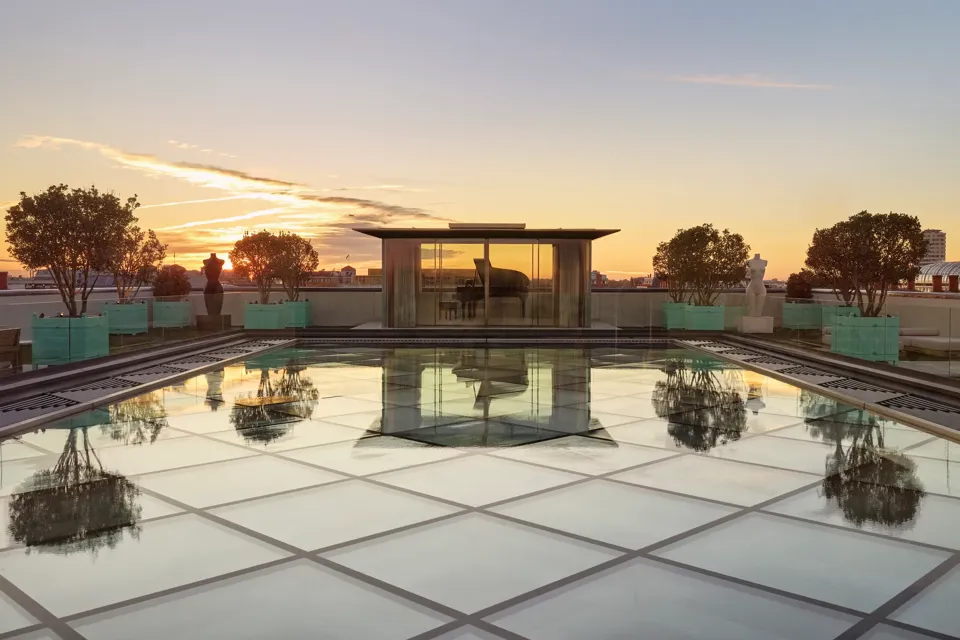 Glass-roof terrace at Claridge’s with a pavilion housing a grand piano, surrounded by trees and sculptures at sunset.
