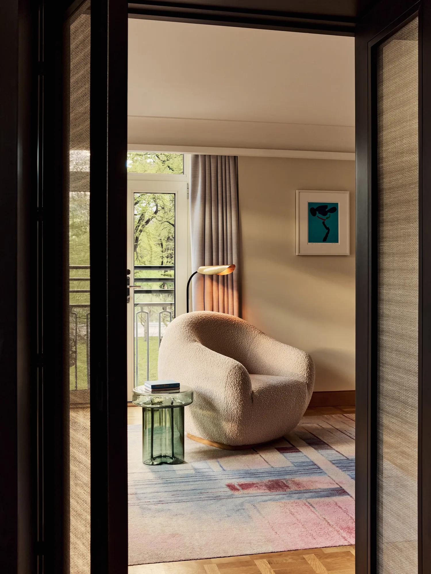 A cosy lounge area featuring a curved white boucle chair, colourful abstract artwork, and a glass side table with books, opening onto a balcony with greenery visible outside