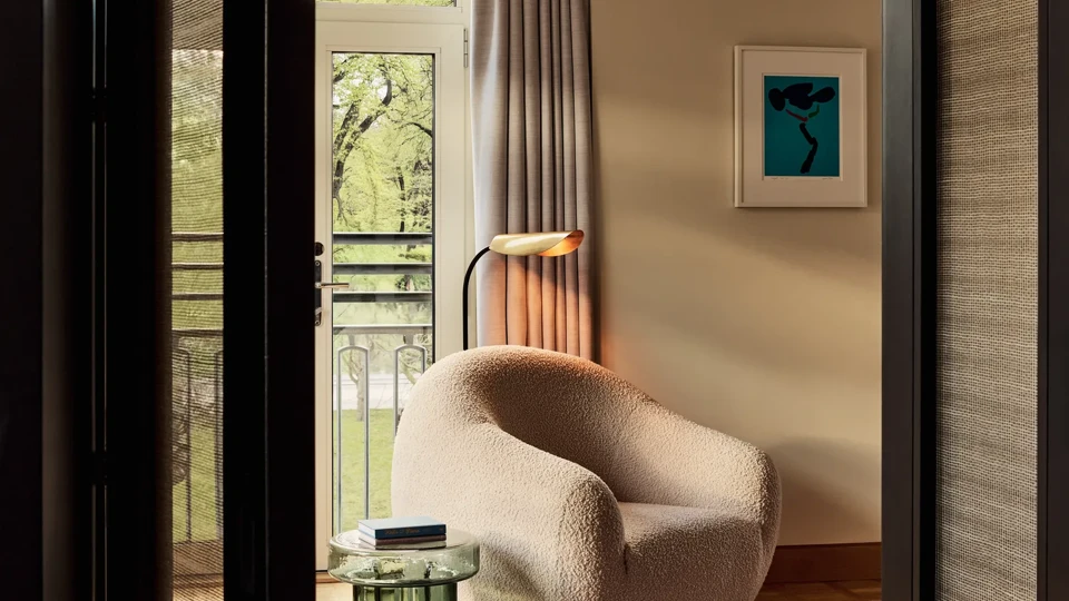 A cosy lounge area featuring a curved white boucle chair, colourful abstract artwork, and a glass side table with books, opening onto a balcony with greenery visible outside