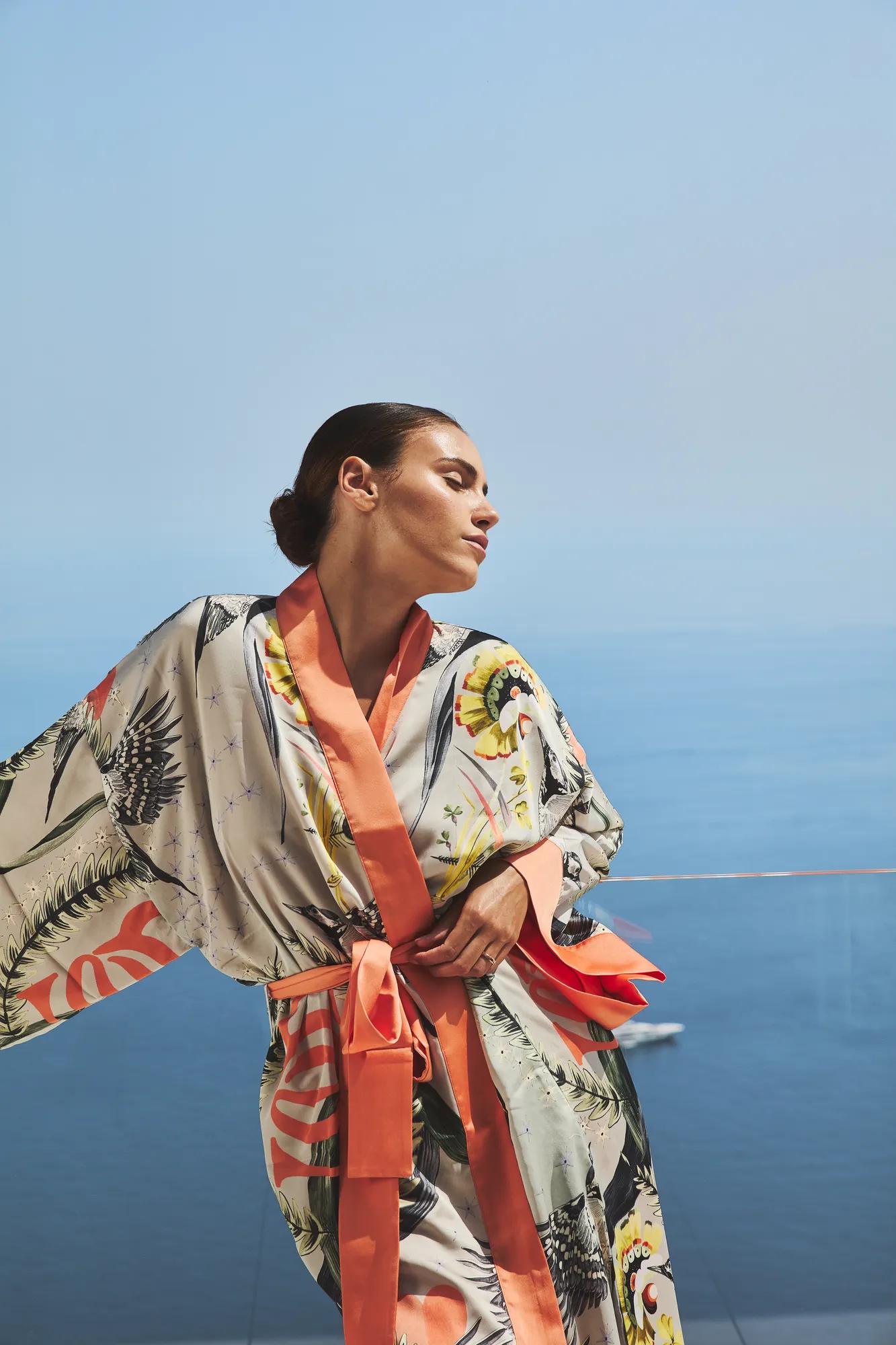 Femme en peignoir de soie imprimé à bordures corail, debout sur une terrasse, les yeux fermés, profitant de la brise marine avec l’océan en arrière-plan.