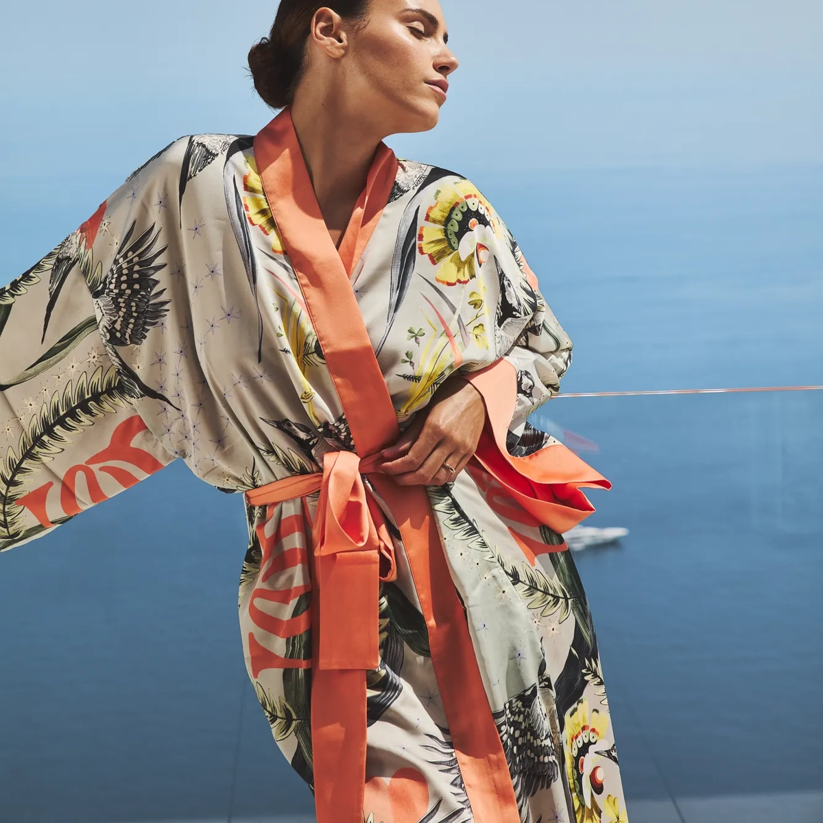 Woman in a patterned silk robe with coral trim standing on a terrace, eyes closed, enjoying the sea breeze with the ocean in the background.