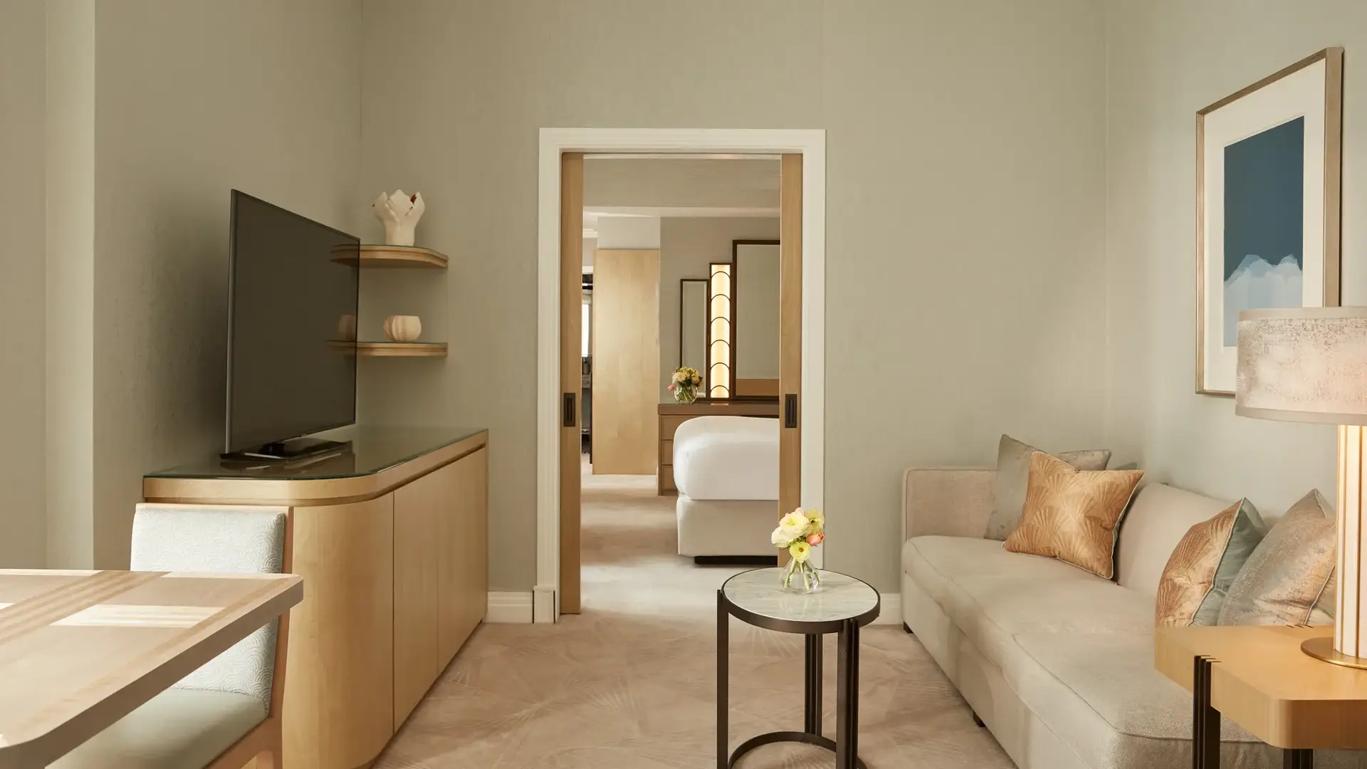 Elegant sitting room with a neutral sofa, side table, and soft lighting, featuring a large TV and dining area, leading through sliding doors to a bedroom with a king bed and warm wooden accents.