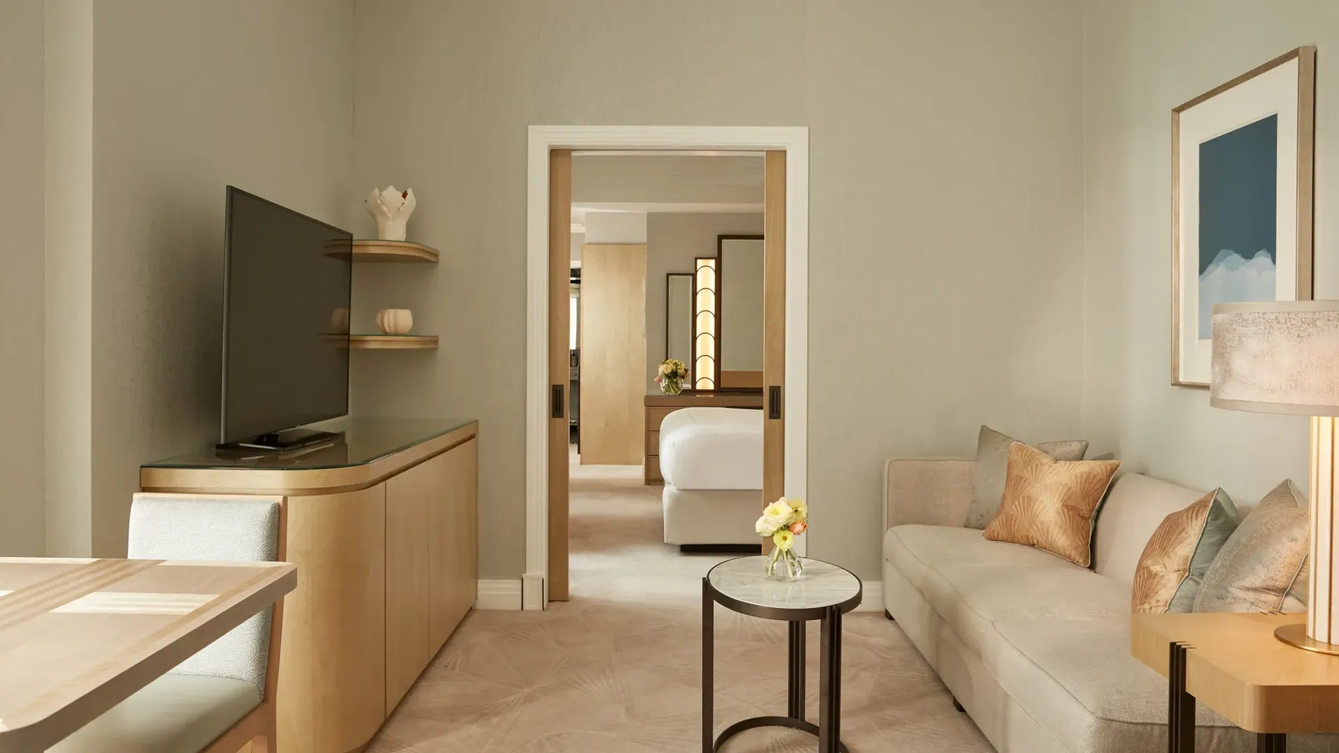 Elegant sitting room with a neutral sofa, side table, and soft lighting, featuring a large TV and dining area, leading through sliding doors to a bedroom with a king bed and warm wooden accents.