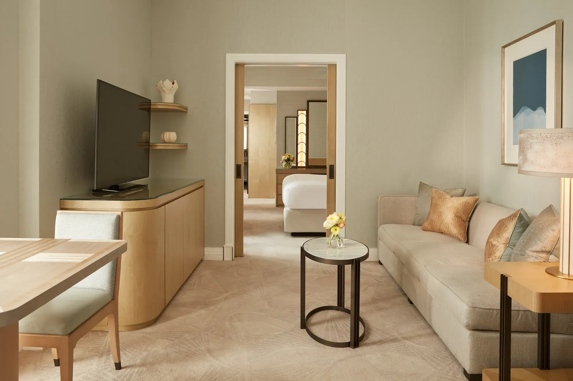 Elegant sitting room with a neutral sofa, side table, and soft lighting, featuring a large TV and dining area, leading through sliding doors to a bedroom with a king bed and warm wooden accents.