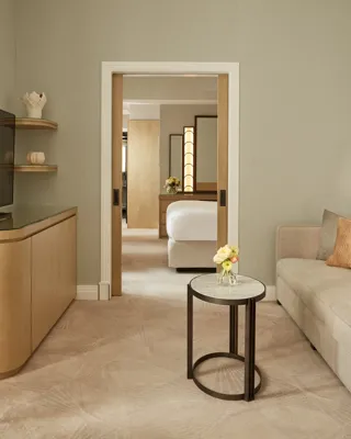 Elegant sitting room with a neutral sofa, side table, and soft lighting, featuring a large TV and dining area, leading through sliding doors to a bedroom with a king bed and warm wooden accents.