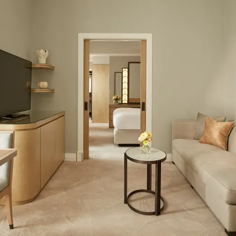 Elegant sitting room with a neutral sofa, side table, and soft lighting, featuring a large TV and dining area, leading through sliding doors to a bedroom with a king bed and warm wooden accents.