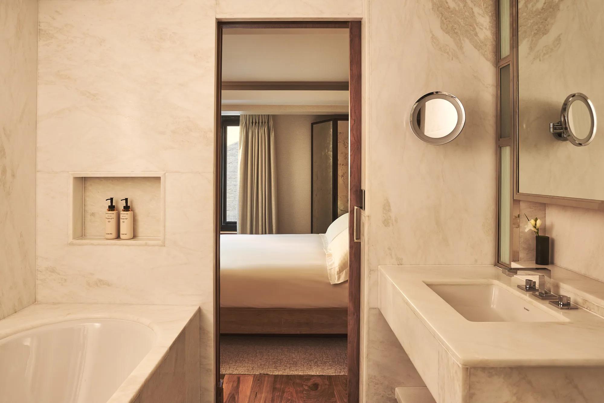 Marble bathroom with a bathtub and vanity, looking through a doorway to a softly lit bedroom with a neatly made bed.