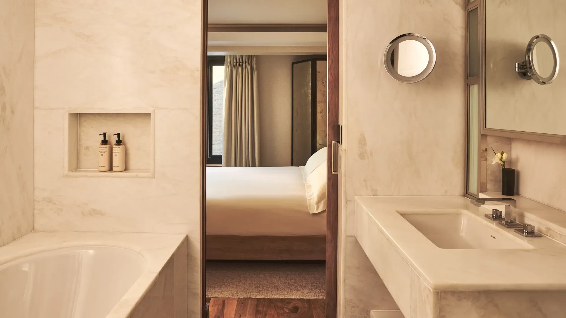 Marble bathroom with a bathtub and vanity, looking through a doorway to a softly lit bedroom with a neatly made bed.
