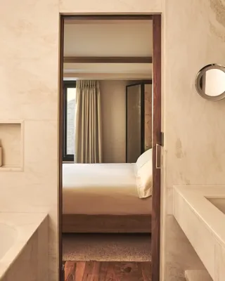 Marble bathroom with a bathtub and vanity, looking through a doorway to a softly lit bedroom with a neatly made bed.