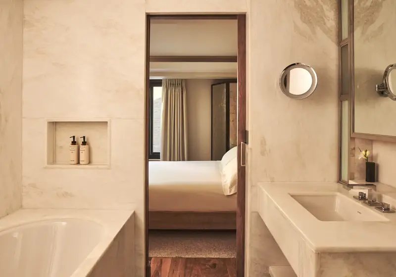 Marble bathroom with a bathtub and vanity, looking through a doorway to a softly lit bedroom with a neatly made bed.